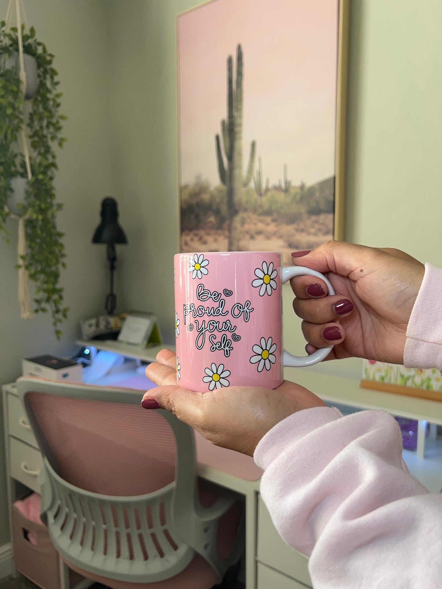 Person holding a pink mug with floral designs and text in a room with a desk and plant.