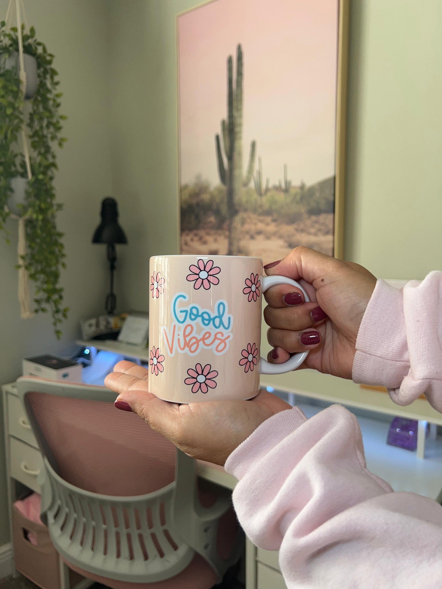 Person holding a mug with 'Good Vibes' text in a room with a cactus painting on the wall.